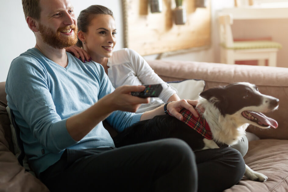 Pareja sonriente junto a su perro viendo tele en el sofá